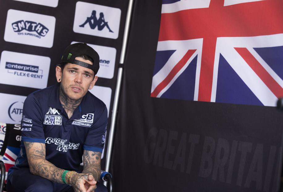 A man in a black Monster Energy cap and a blue polo shirt with "GB Speedway Team" and sponsor logos, sits in front of a Union Jack flag and a banner with "GREAT BRITAIN" and various brand logos.