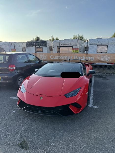 A red Lamborghini supercar parked in a lot, with a banner for "Customer Parking" and "Motorhome Holiday Company" in the background.