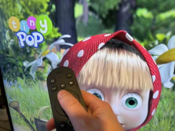 A hand holding a Sky TV remote, pressing a button while the animated show "Tiny Pop" is displayed.