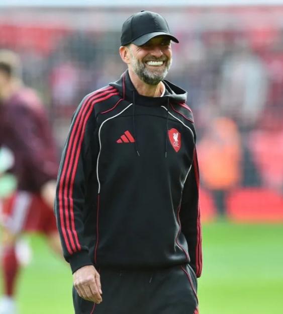 Jürgen Klopp smiles while wearing a black Liverpool F.C. hoodie and a black cap.