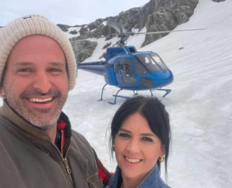 A man and woman smiling for a selfie with a helicopter in the background on a snowy mountain.