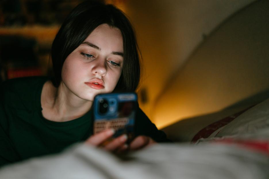 A teenage girl lying in bed and looking at her mobile phone in the dark, with the light from the phone illuminating her face.