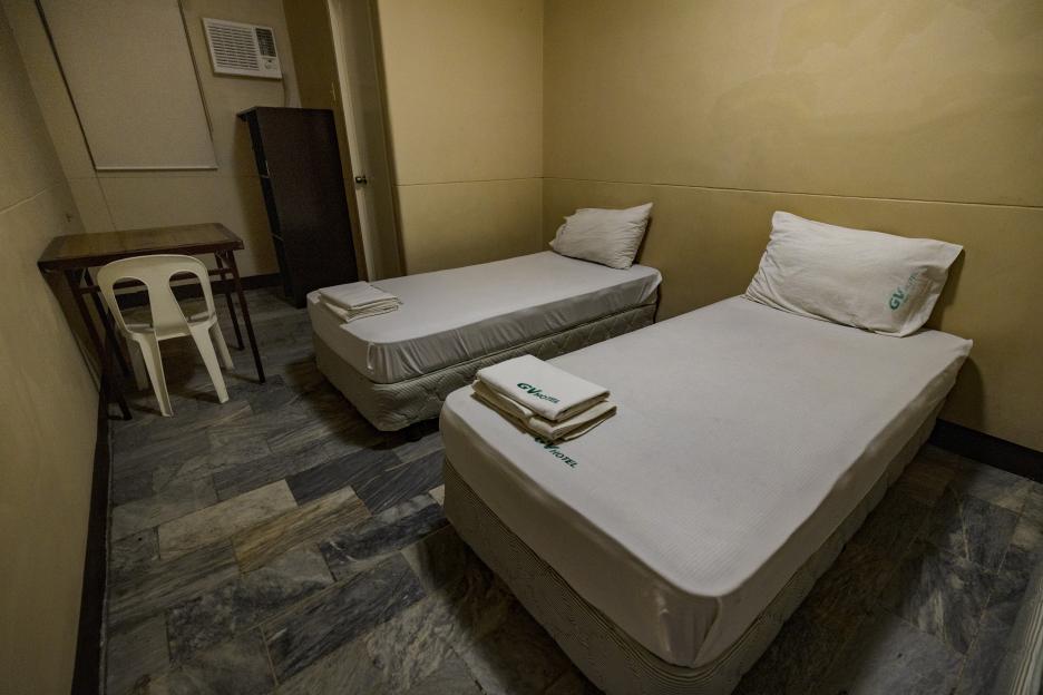 A GV Hotel room with two single beds, a small table, a white chair, and an air conditioner.