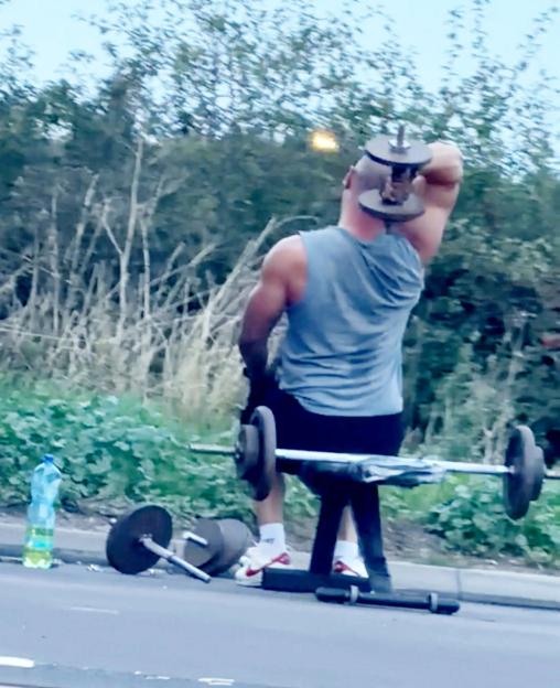 A lorry driver doing a triceps extension with a dumbbell on the hard shoulder of a road.