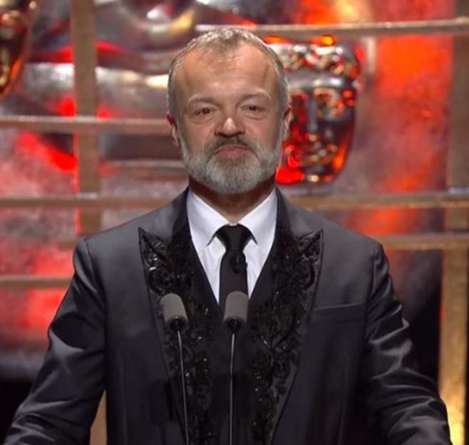 Graham Norton speaking at a podium with two microphones, wearing a black suit with embroidered lapels.