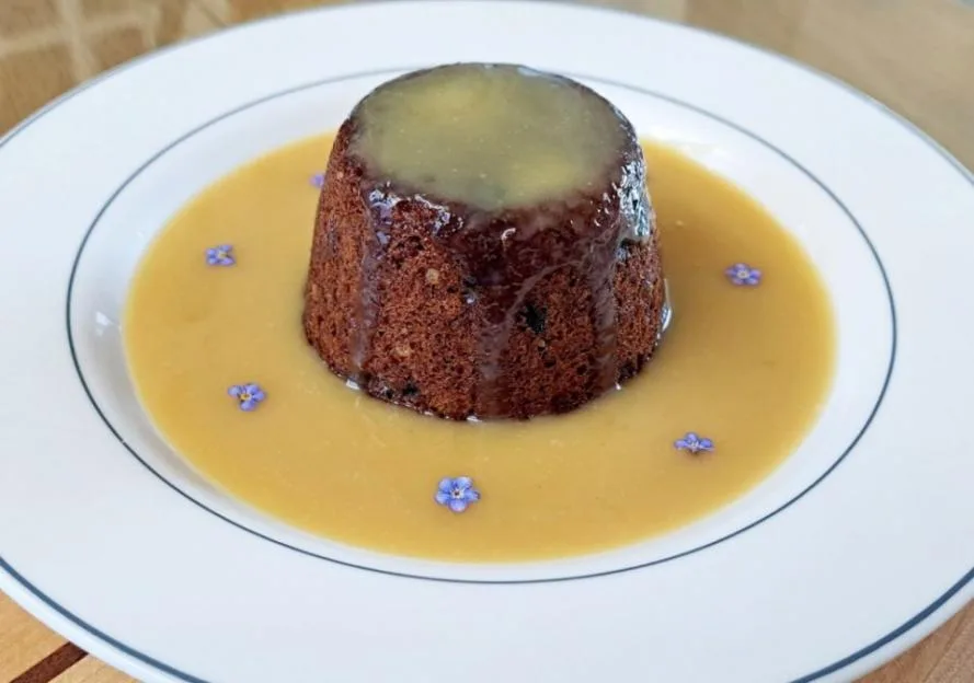 A steamed sponge pudding in a pool of custard on a white plate.