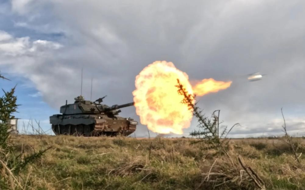 A tank firing a projectile with a large muzzle flash, seen from behind a bush.