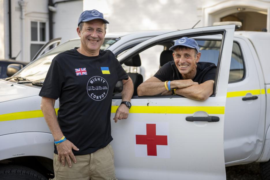 Two men, wearing Mighty Convoy hats and shirts with the UK and Ukrainian flags, smile next to an ambulance with a red cross.