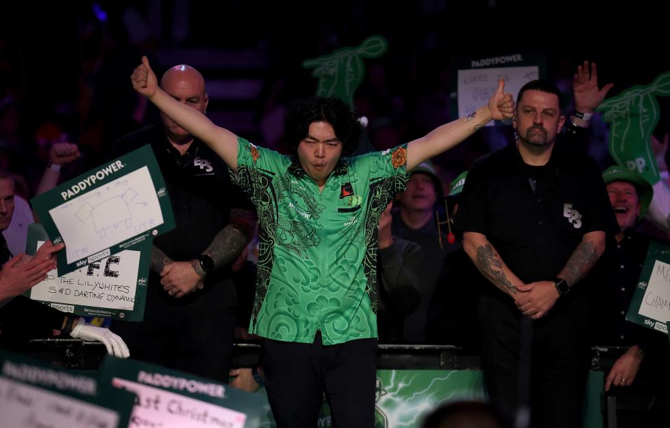 Motomu Sakai of Japan walks onto the stage with his arms raised, wearing a green shirt, at the 2025/26 Paddy Power World Darts Championship.