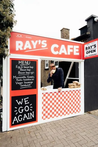 A man eating a sandwich at Ray's Cafe, a popup cafe with a red and white sign, menu, and checkered counter.