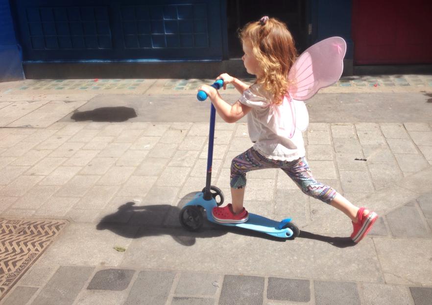 UK, England, London, Portrait of girl (2-3) with fairy wings riding scooter on city pavement