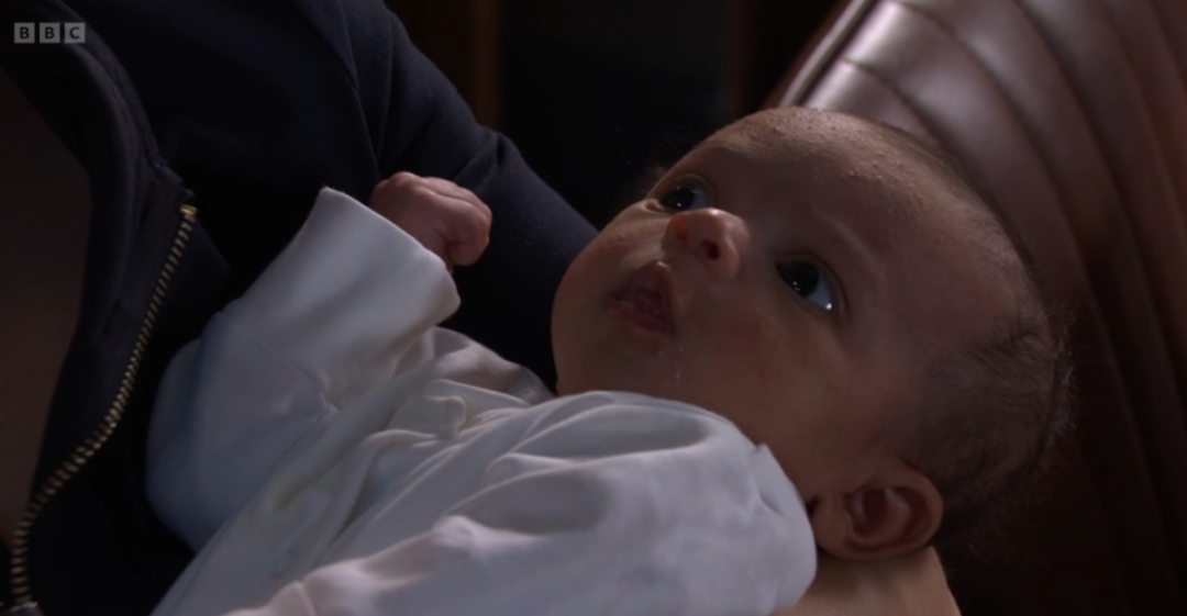 A baby with dark hair, looking up, with a BBC logo visible in the top left corner.