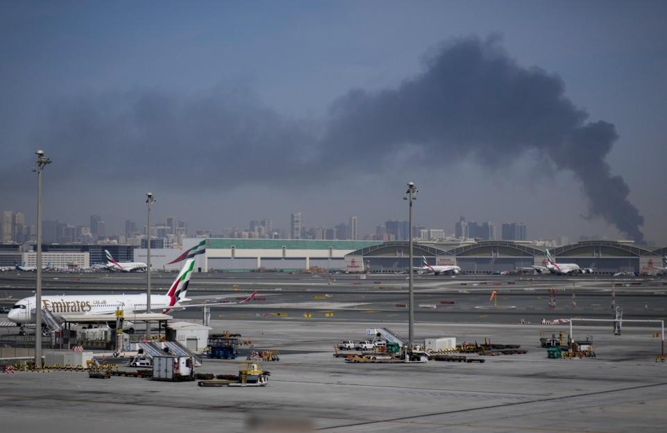 Plumes of dark smoke rising in the distance from an airport in front of a city skyline.