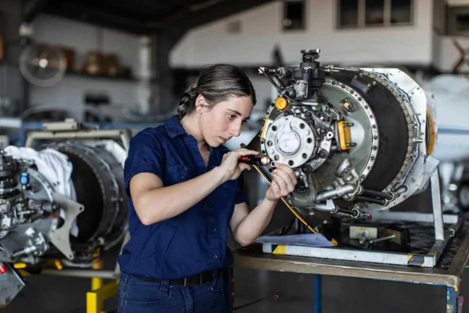 A young female aircraft engineer apprentice inspecting an aircraft engine with a flashlight in a hangar.