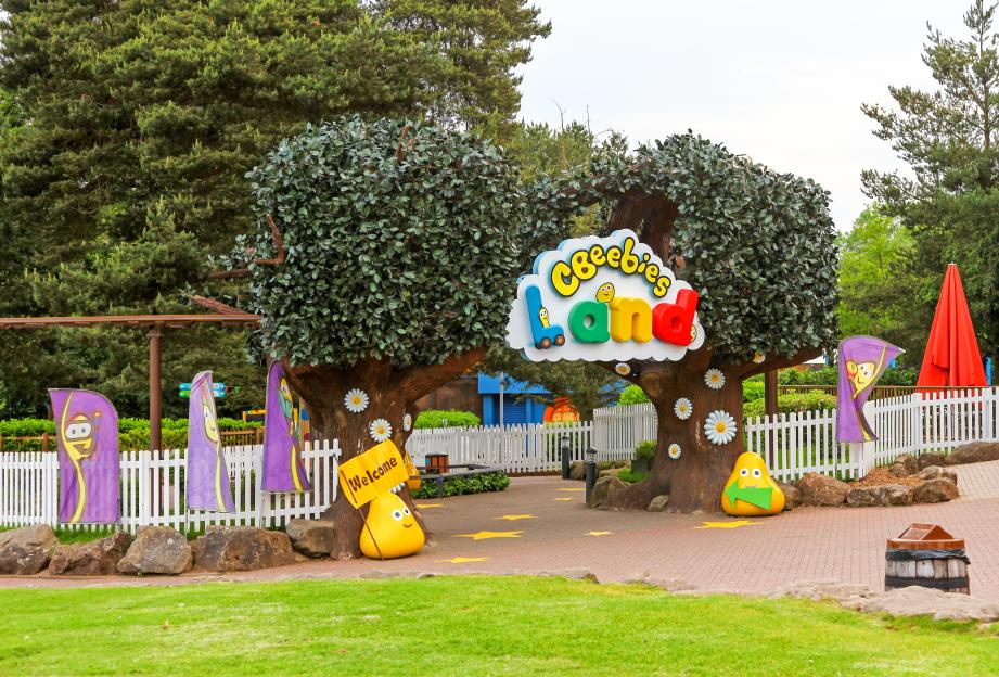 Entrance to Cbeebies Land at Alton Towers, with a large colorful sign for "Cbeebies Land" between two large green topiary trees with white daisies on their trunks.
