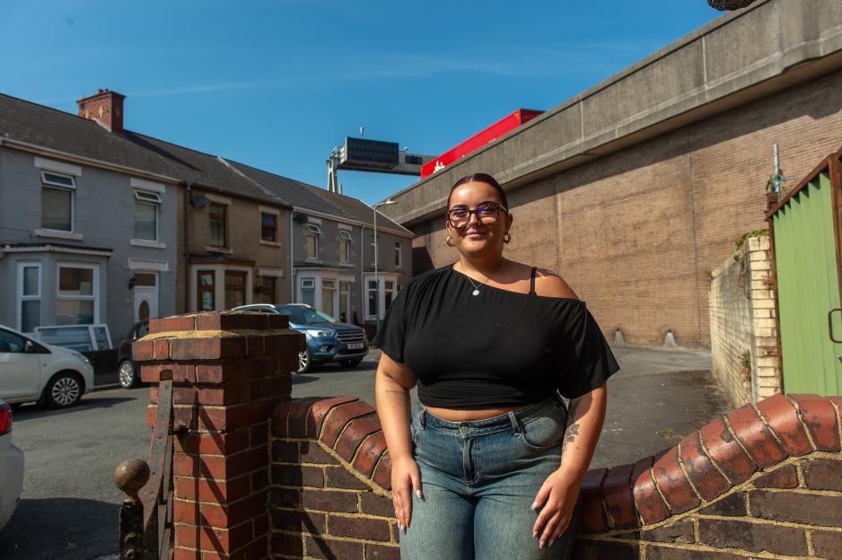 Evie Cursons, a woman in glasses, stands smiling in front of a brick wall, with homes and an overhead motorway behind her.