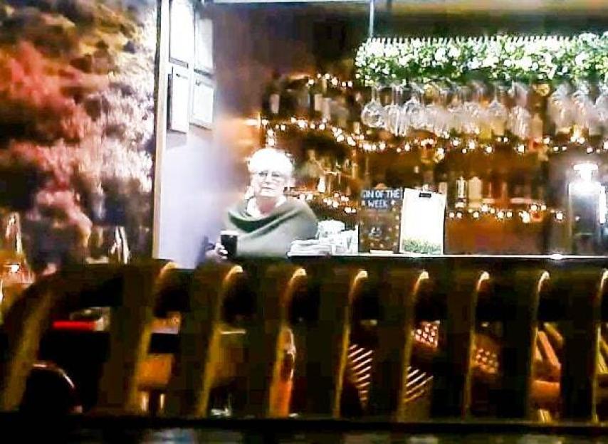A woman wearing a green shawl stands behind a bar holding a drink.