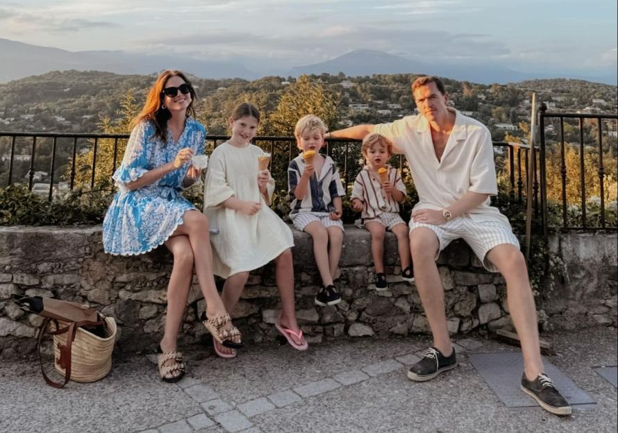 A family of five, including a mother, father, and three children, sitting on a stone wall overlooking a scenic mountain landscape, all holding ice cream cones.