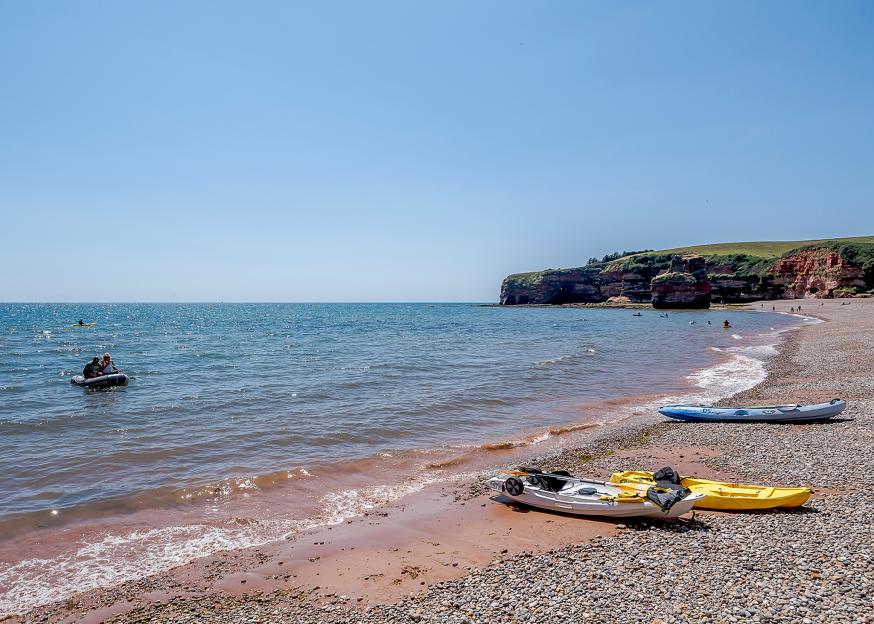 Ladram Bay Holiday Park in Devon, with boats on the water and kayaks on the pebble and sand beach.