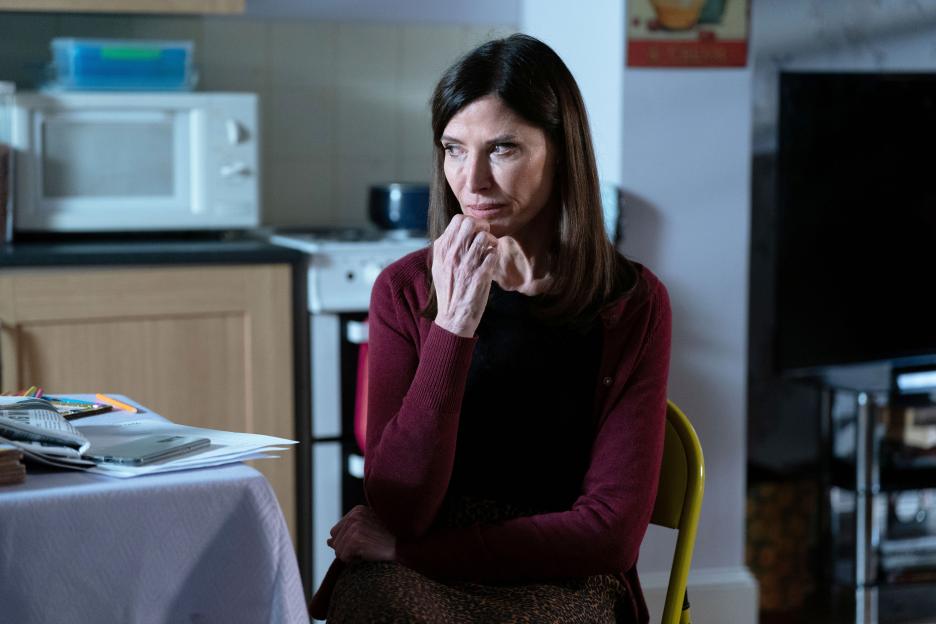Bea (Ronni Ancona) sits at a table with papers and a phone, looking thoughtful.