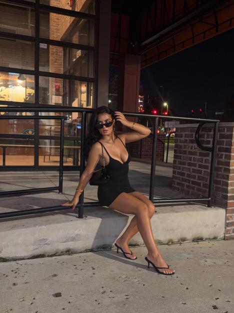 A woman wearing a black mini dress and sunglasses sitting on a ledge.