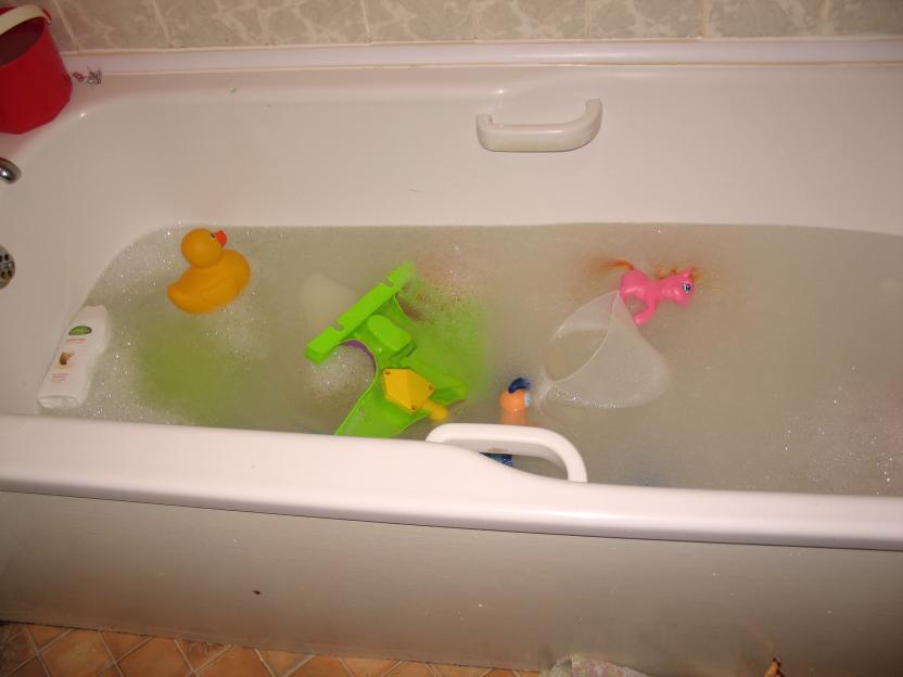 Bubble bath with a rubber duck, green toy, pink unicorn, and clear measuring cup floating in it.