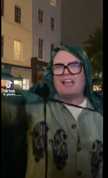 Person in glasses wearing a green hooded cape and light green patterned shirt, standing in front of buildings.