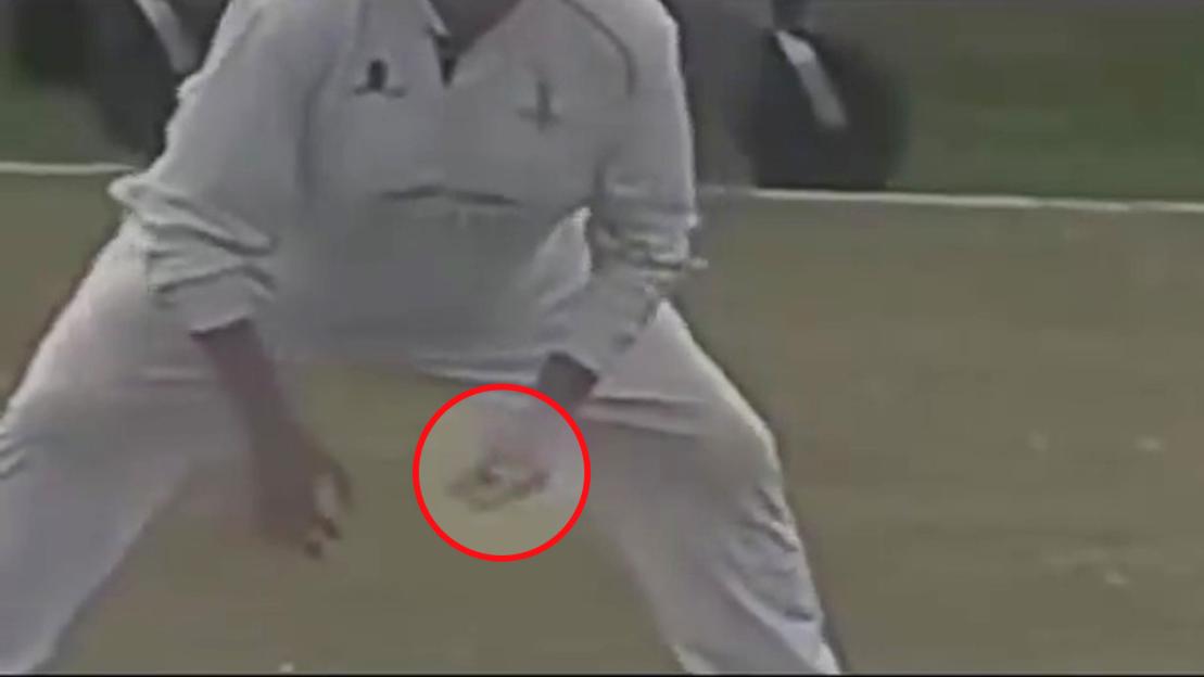 A cricket player in white uniform holding something small in his hand, highlighted by a red circle, on a field during a game.
