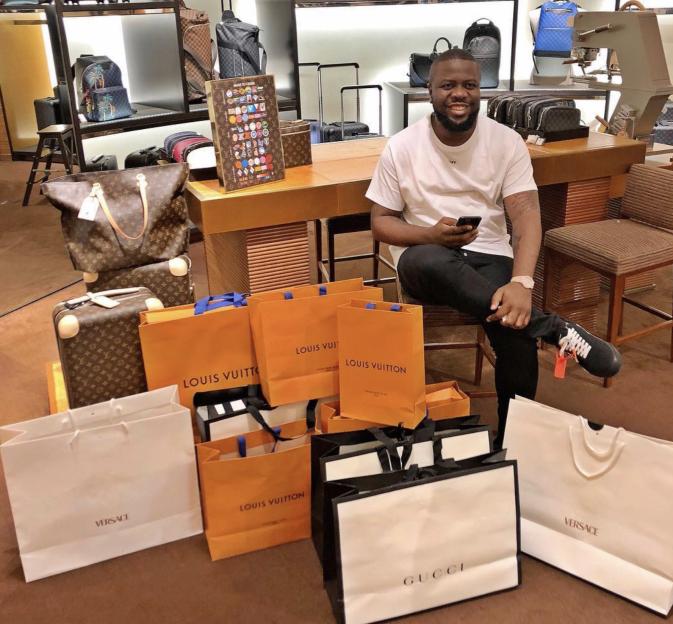 Ramon Abbas, known as Ray Hushpuppi, sitting with luxury shopping bags from brands like Louis Vuitton, Versace, and Gucci surrounding him.