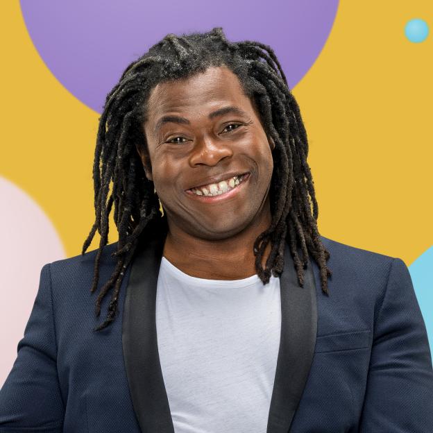 Ade Adepitan MBE smiling at the camera, wearing a white t-shirt and blue jacket.