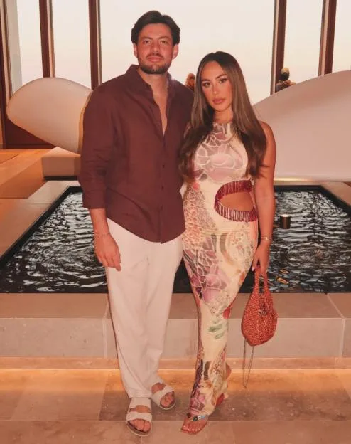 A man and woman standing together in front of a water feature, with the man wearing a brown shirt and white trousers and the woman in a patterned, cut-out dress holding a red bag.