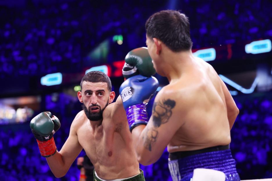 Two boxers in a ring during a fight, one with a beard and green gloves, the other with blue gloves and tattoos.