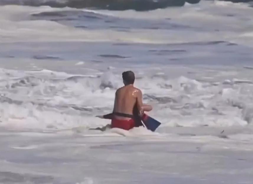 A person in red shorts and a dark rescue tube sits in crashing waves, looking out at the ocean.