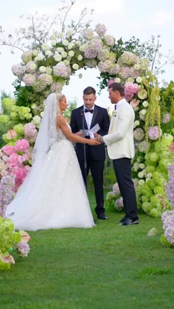 A couple at an outdoor wedding ceremony, exchanging vows in front of an officiant under a floral arch.