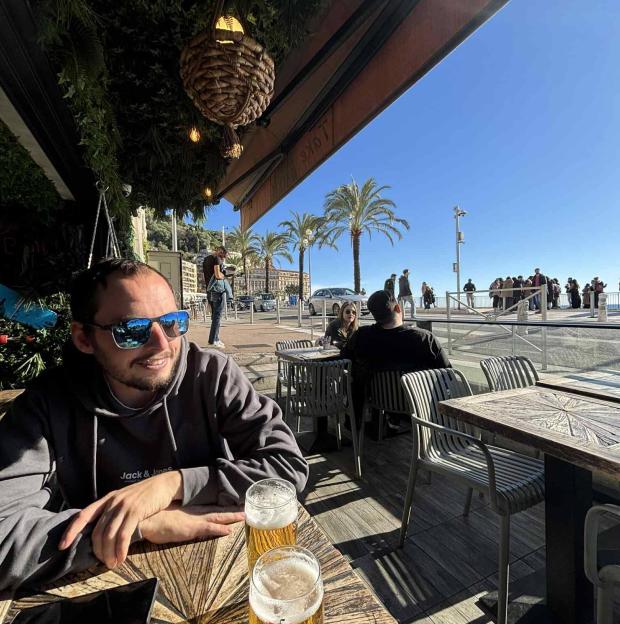 A man in sunglasses and a hoodie sits at an outdoor table with two beers, facing a sunny street with palm trees, people, and buildings.