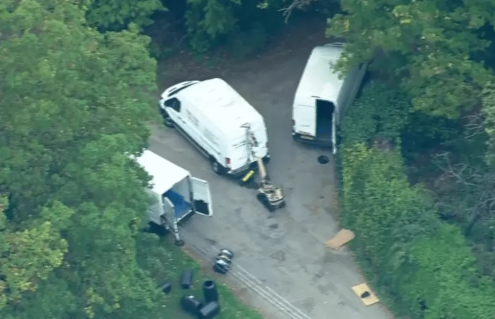 Three white vans and an EOD robot on a paved area surrounded by trees.