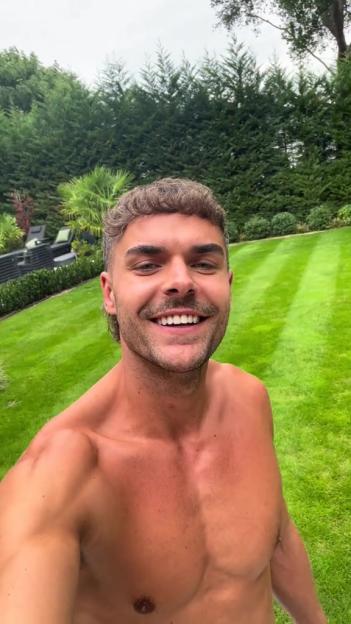 A smiling shirtless man with a mustache stands in a large, well-manicured garden.
