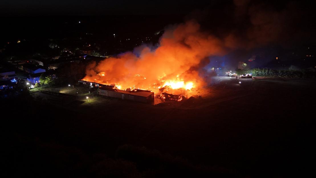 A large building engulfed in flames at night, with bright orange fire and smoke rising into the dark sky, and fire trucks and personnel visible on the right.