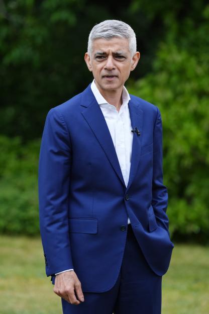 Sadiq Khan, Mayor of London, wearing a blue suit and white shirt, outdoors in front of greenery.