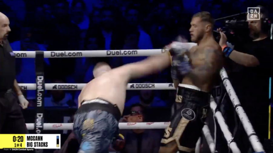 A boxer, McCann, in white and blue shorts and no shirt, throws a right hook at an opponent in black and gold shorts.