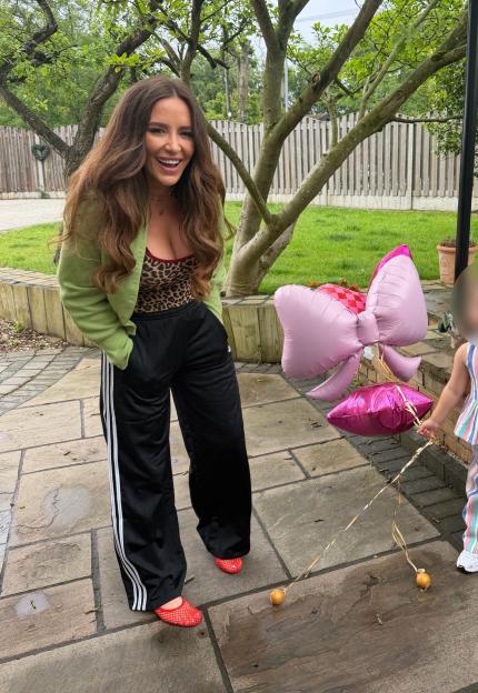 A woman smiles at the camera, dressed in a green cardigan, leopard print top, black track pants, and red mesh shoes, holding balloons next to a child.