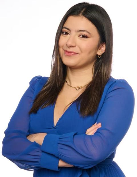A woman with long dark hair, wearing a royal blue dress and gold jewelry, smiles with her arms crossed.