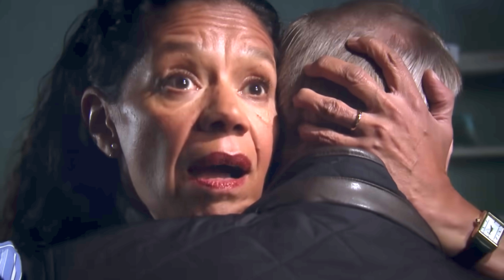 A woman with dark hair and dark red lips, with wide-open eyes and mouth, embraces a man with gray hair, his back to the viewer.