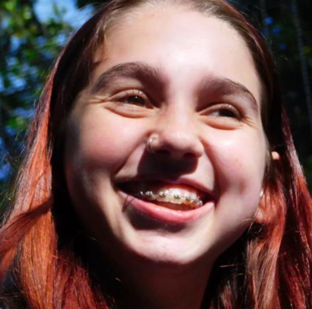 Portrait of Ellame Ford-Dunn, a smiling young woman with red hair and braces, looking up and to the right.