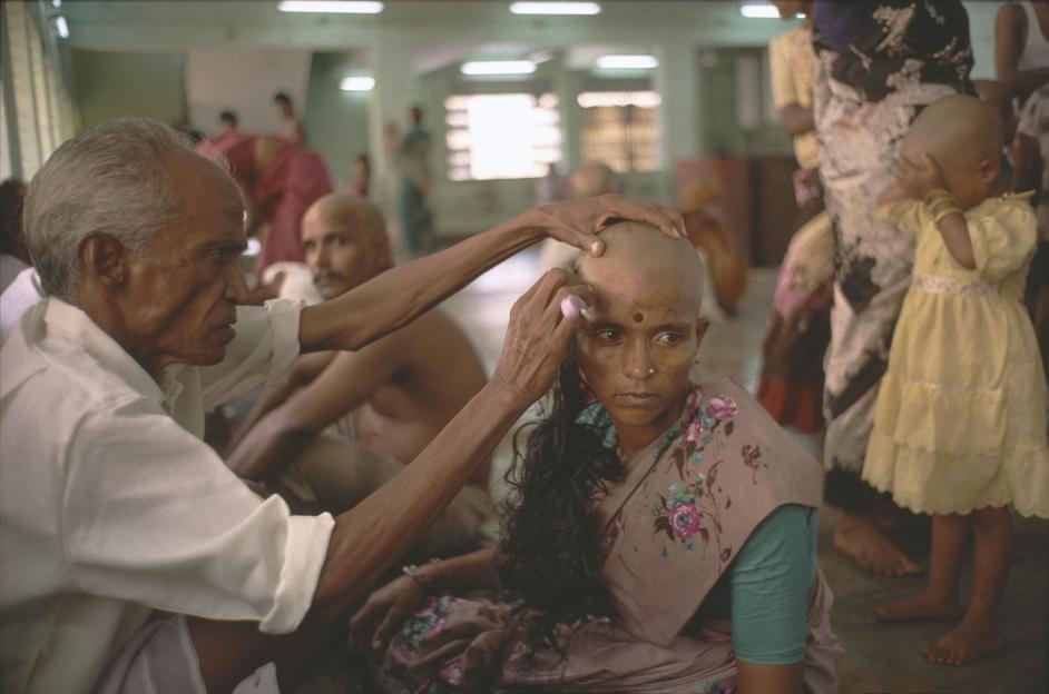 A man shaves a woman's head in the Tirumala temple, with other people in the background, including a small child covering their eyes.