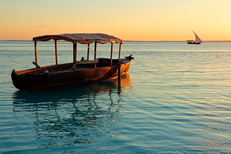 A wooden boat with a canopy floating on calm turquoise water at sunset, with a sailing boat in the distance.