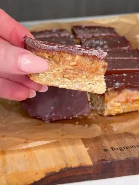A hand holding a chocolate-covered caramel and oat bar, with more bars on parchment paper on a wooden cutting board.