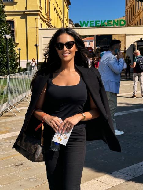 A woman in a black top, pants, and blazer, wearing sunglasses and holding a water bottle and papers, smiles in front of a building with a "WEEKEND" sign.
