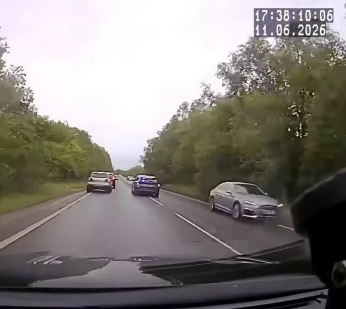 Dashcam view of a grey car overtaking on a two-lane road with a forested median.