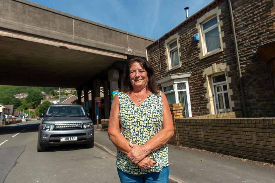 A woman standing in front of a stone house with a highway bridge overhead and a car parked on the street.